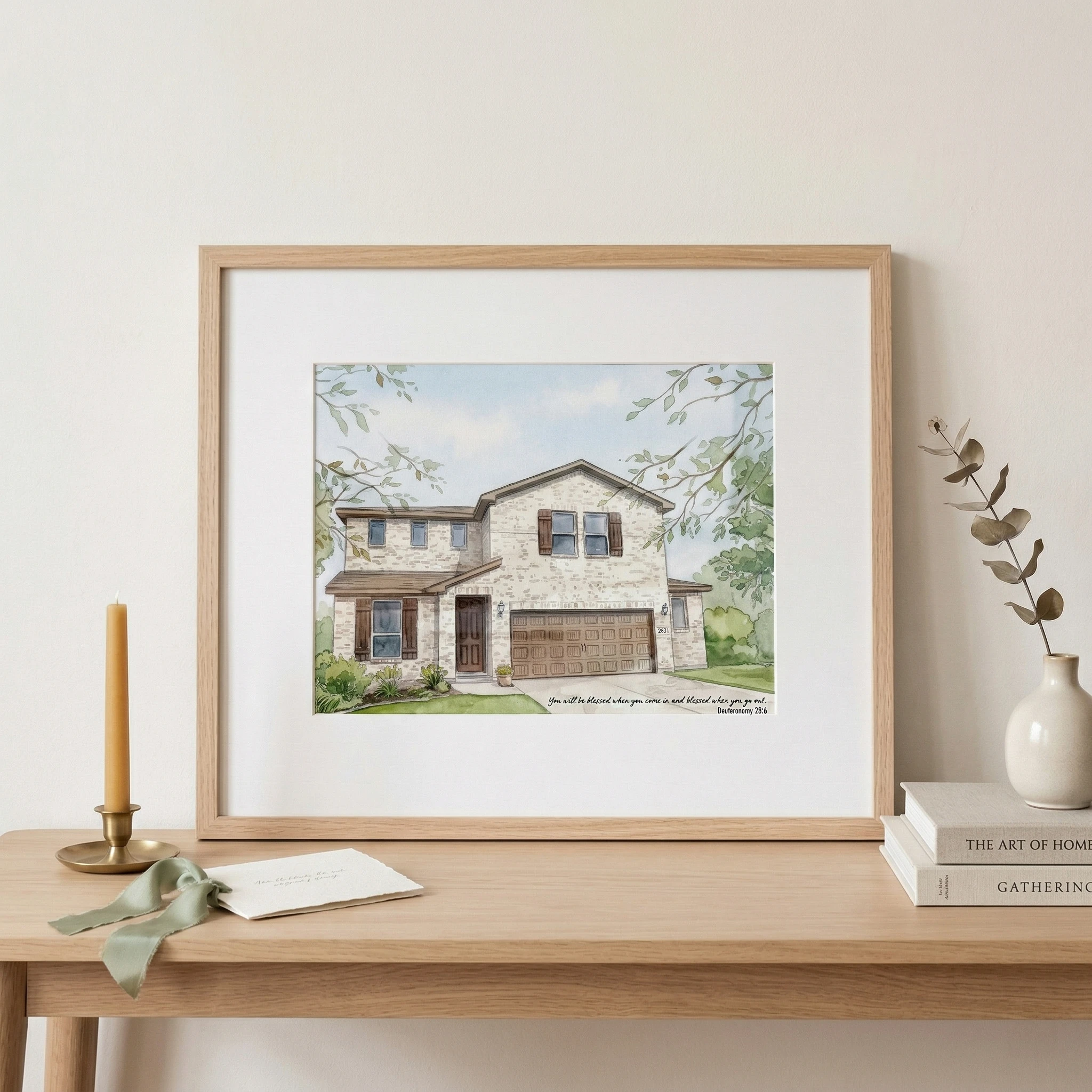 Framed house portrait displayed on a styled console table with a note card and greenery.